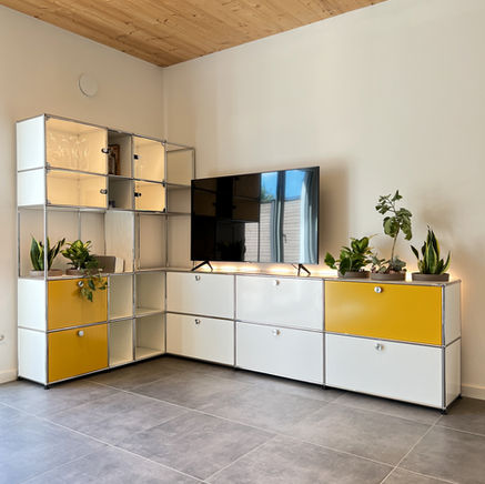 Großes L-förmiges Modularregal von USM-Haller in Weiß und Gelb mit Glaselementen, integriertem Fernseher und Grünpflanzen auf hellem Fliesenboden.