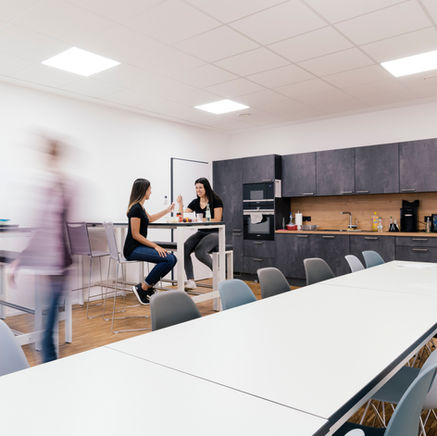 Helle Büroküche mit zwei Mitarbeiterinnen am Stehtisch, dunkler Einbauküche und weiteren Sitzgelegenheiten an weißen Tischen.