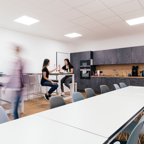 Helle Büroküche mit zwei Mitarbeiterinnen am Stehtisch, dunkler Einbauküche und weiteren Sitzgelegenheiten an weißen Tischen.