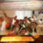 Group of people in beige suits celebrate, posing energetically in an office. Whiteboard message and "Karla Rensburg" nameplate are visible.