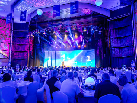Wide view of an indoor event venue with seated guests, a large stage screen and purple-blue spotlights, photographed by an event photographer in Budapest.