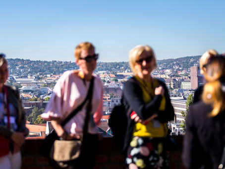 Rendezvény résztvevői egy budapesti panorámateraszon: az előtérben szándékosan elmosódó, napszemüveges vendégek, a háttérben éles városkép és domboldal.