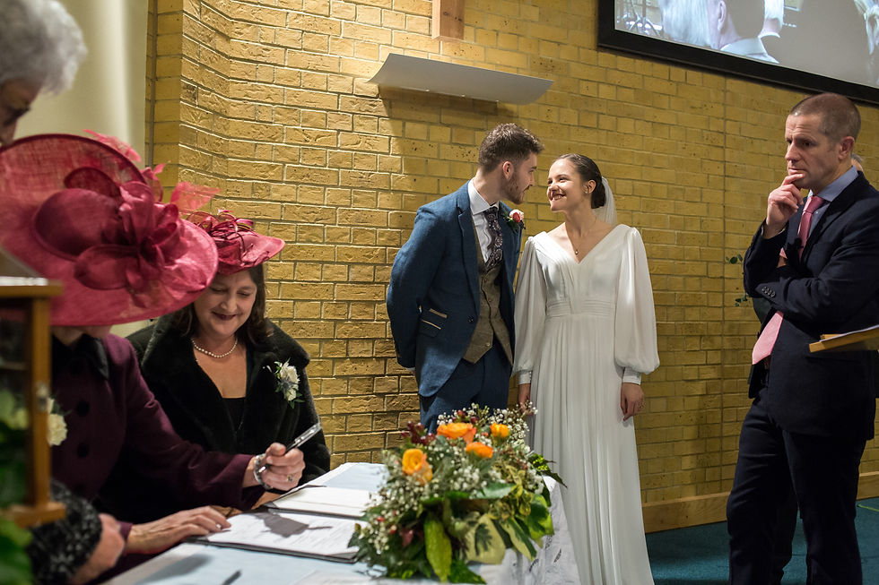 Bride and groom smiling in a ceremony with a brick wall background. Two women in red hats sign documents, and a man observes thoughtfully.