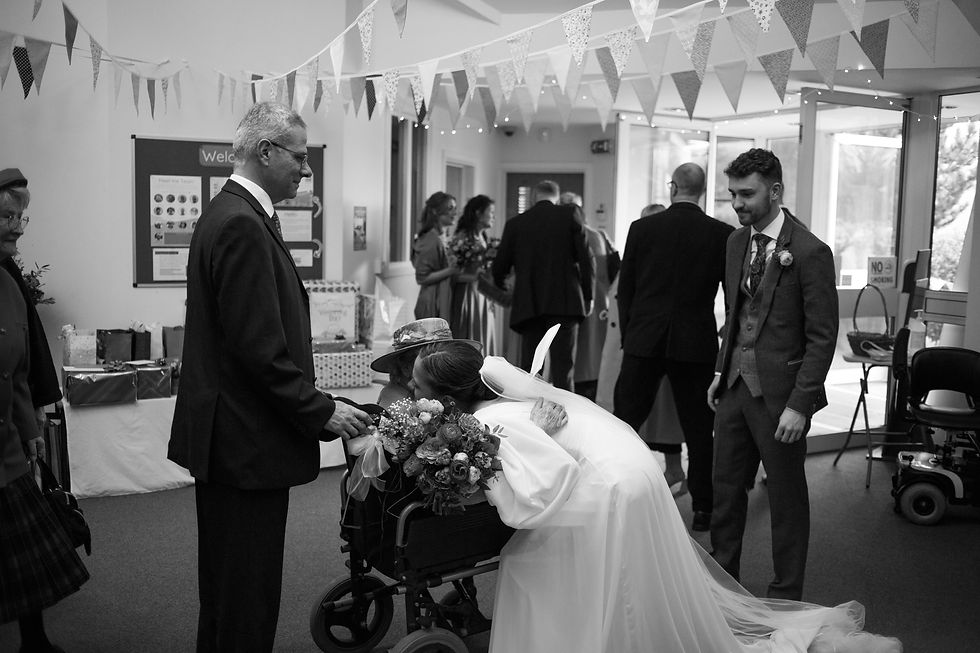 Bride hugs an elderly woman in a wheelchair, holding a bouquet. Guests are gathered under bunting in a venue, creating a warm atmosphere.