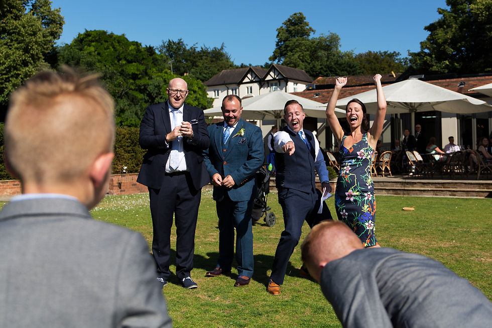 Wedding guests cheer and laugh outdoors. A woman raises her arms in victory. Sunny day, house and patio in the background.