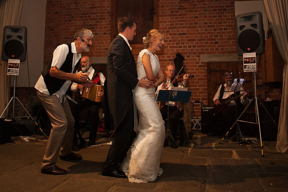 Bride and groom dance joyfully indoors with a band playing accordion and violin in the background, set against a red brick wall.