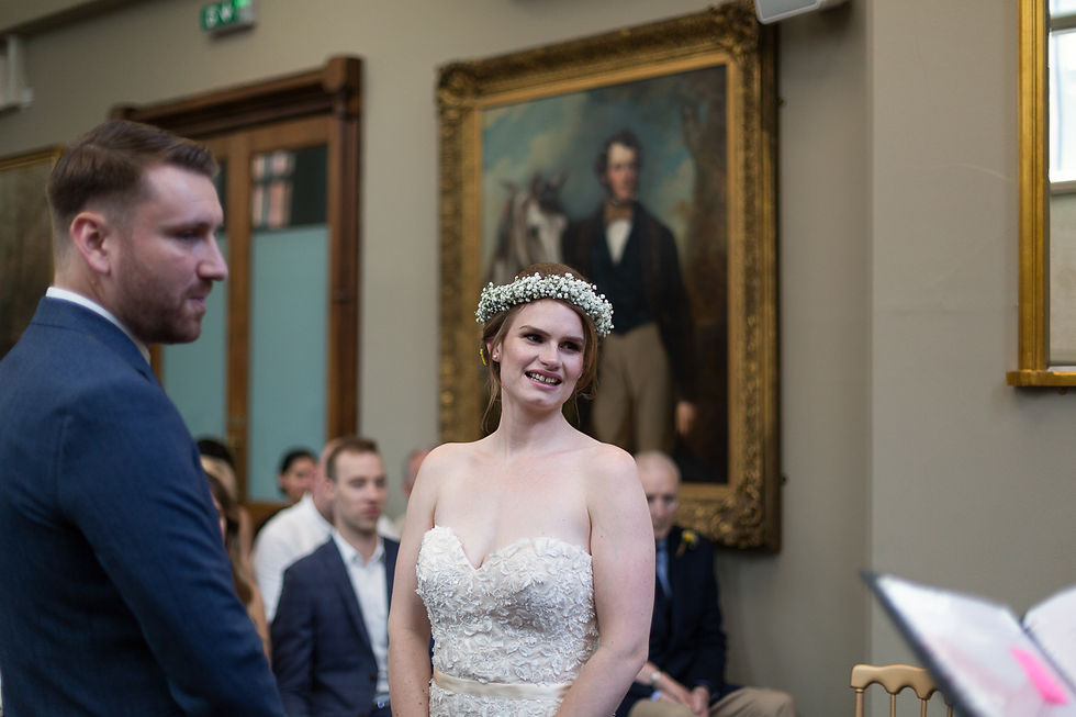 Bride in lace dress smiles, wearing a floral crown. Groom gazes at her. Formal room with a portrait in the background. Guests seated.