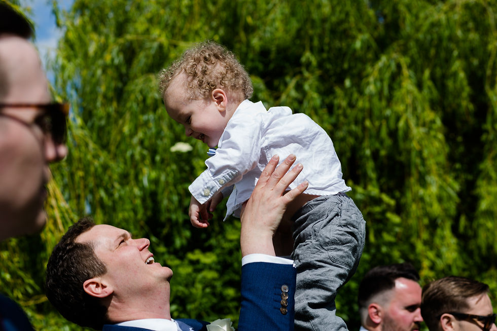Man in blue suit lifting smiling toddler in white shirt. Green, leafy background. Others in foreground. Joyful outdoor setting.