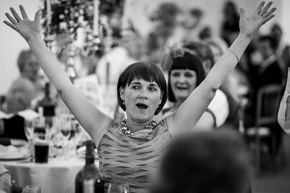 Woman with arms raised, smiling in excitement at a formal event. Black and white, blurred guests in background, festive atmosphere.