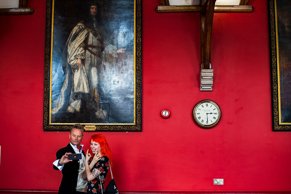 Two people taking a selfie in a red room. A large portrait and a clock are on the wall. The woman has bright red hair and tattoos.