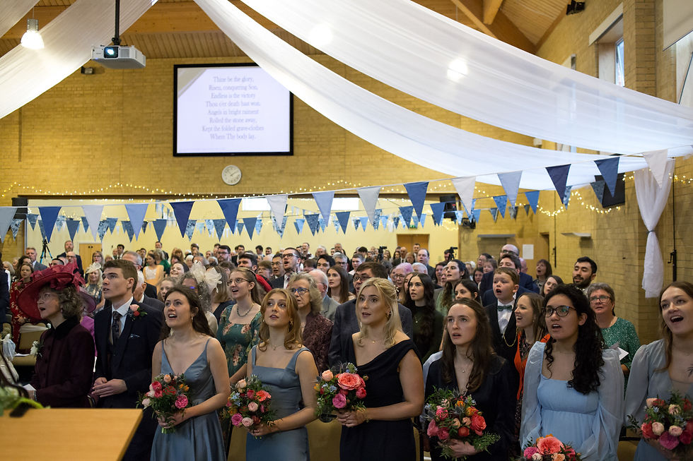 People dressed formally singing in a decorated hall with blue and white pennants. Flowers in hand. Projected hymn lyrics in background.