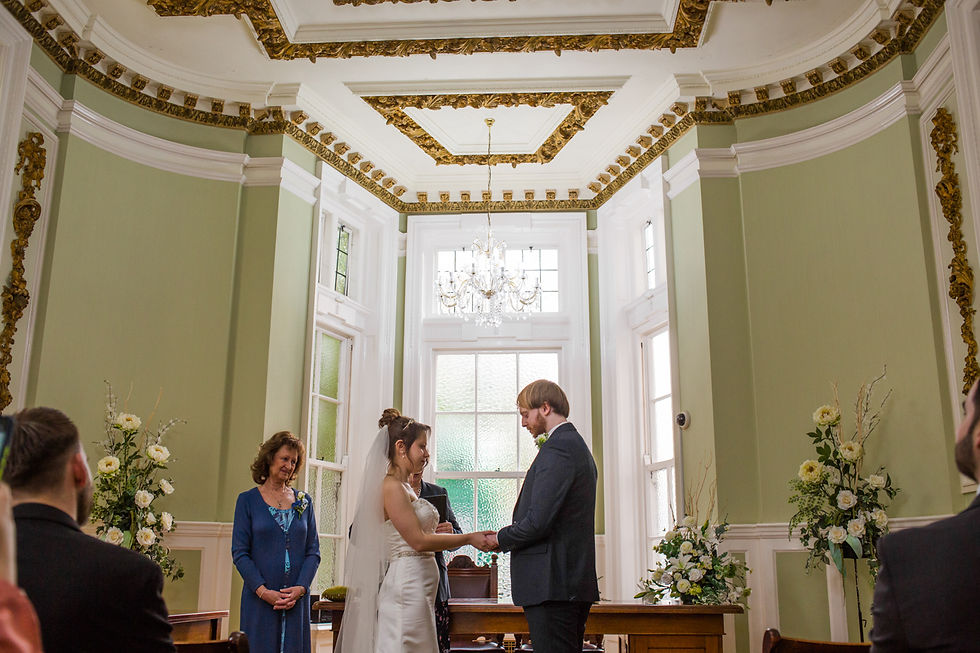 Bride and groom exchange vows in an elegant room with green walls and gold accents. A woman in blue stands nearby. Flowers decorate the space.