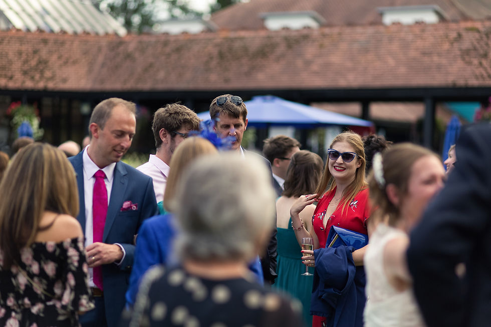 People at an outdoor event, mingling. A woman in red with sunglasses smiles, holding a glass. Brick building and blue umbrellas in background.