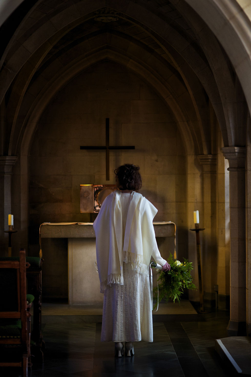 Bride in white, holding greenery, stands in a dimly lit chapel facing an altar with a cross. Moody atmosphere with candles lit.