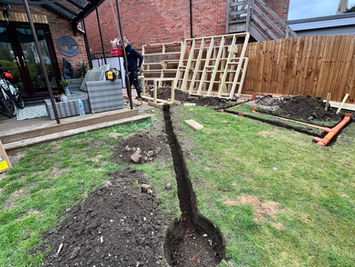 Attgills Drainage Services - Annex Drainage Northamptonshire - Narrow drainage trench dug across a lawn leading to a circular pit, with soil piles and timber framework visible in the background.