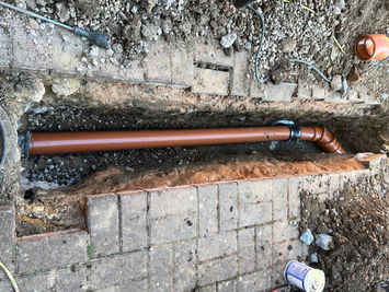Attgills Drainage Services - Annex Drainage Northamptonshire - Trench under block paving with newly installed orange PVC drainage pipe on gravel bed, connected by a black coupling to an angled fitting.