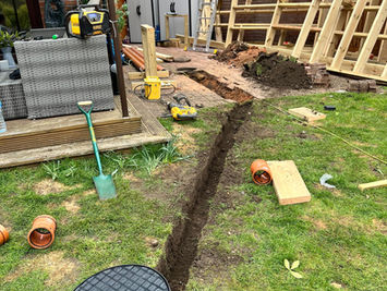 Attgills Drainage Services - Annex Drainage Northamptonshire - Narrow garden trench leading to an inspection chamber, with drainage pipes, timber framework, and tools arranged around the work area.