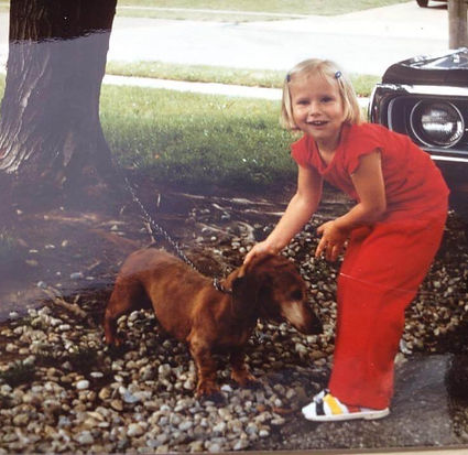 A little girl in a red outfit pets a red dachshund in a driveway in 1976.