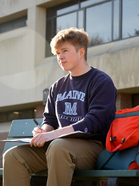 A University of Maine student sitting on a campus bench, writing notes in a notebook.