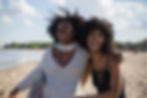 two black girls laughing alongside the coastside of a beach smiling and hugging one another