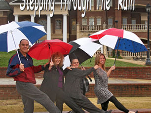 4 people walking in rain with umbrellas with a hand on shoulder of person in front of them, and each with right foot stepping forward. Text on image is "Stepping through the rain"(Original photography Sheri & Bill)