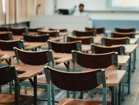 Empty classroom with rows of wooden chairs and desks. A person is blurred in the background, standing at a lectern, suggesting a lecture setting.