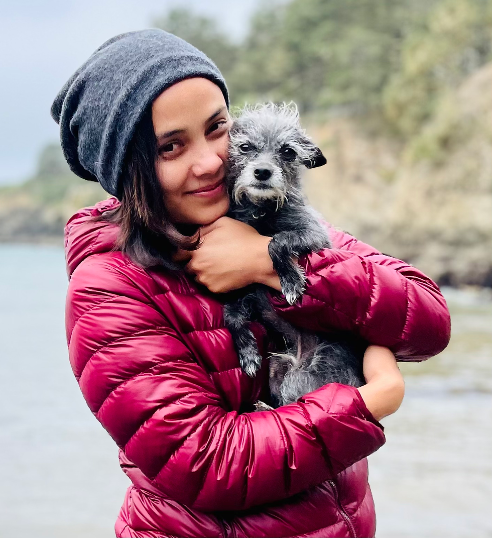 Woman in a red jacket (Raquel Bernaldo, from FAAS) smiles tenderly and hugs a small gray dog by a lake. She's wearing a gray beanie. The background is blurred greenery, evoking warmth.