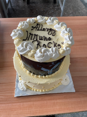 Yellow and white frosted cake with the words ‘Allergy Immuno Rocks’ written on top, placed on a classroom desk