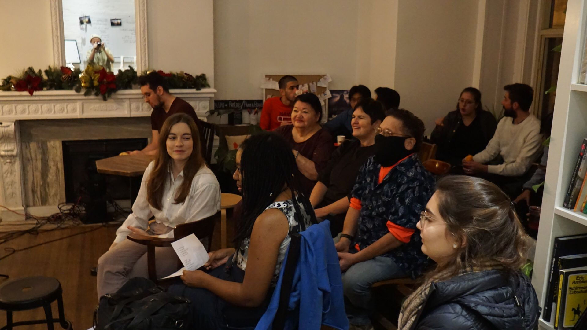 A seated audience watches the performance, with some chatting and smiling in a warmly decorated room.