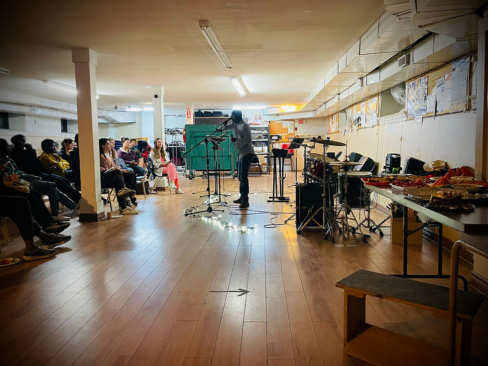 Ernest Okyere-Twum speaks into a microphone in a warmly lit basement venue, with a seated audience on the left and instruments and food setup on the right.