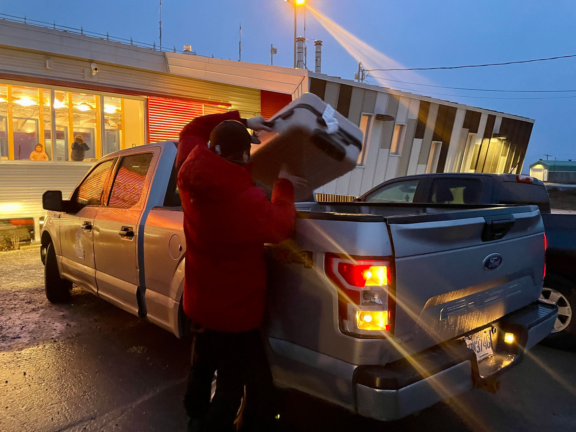 a man loading a grey suitcase into a pickup truck outside the Kuujjuaq airport at dusk
