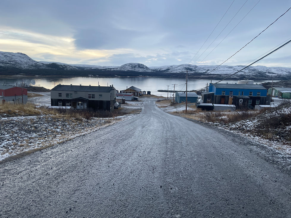 Alt text: “View down a sloping gravel road toward the water in Tasiujaq, Nunavik. Snow dusts the ground and rooftops of small houses under a grey sky, with low mountains and a calm inlet in the distance