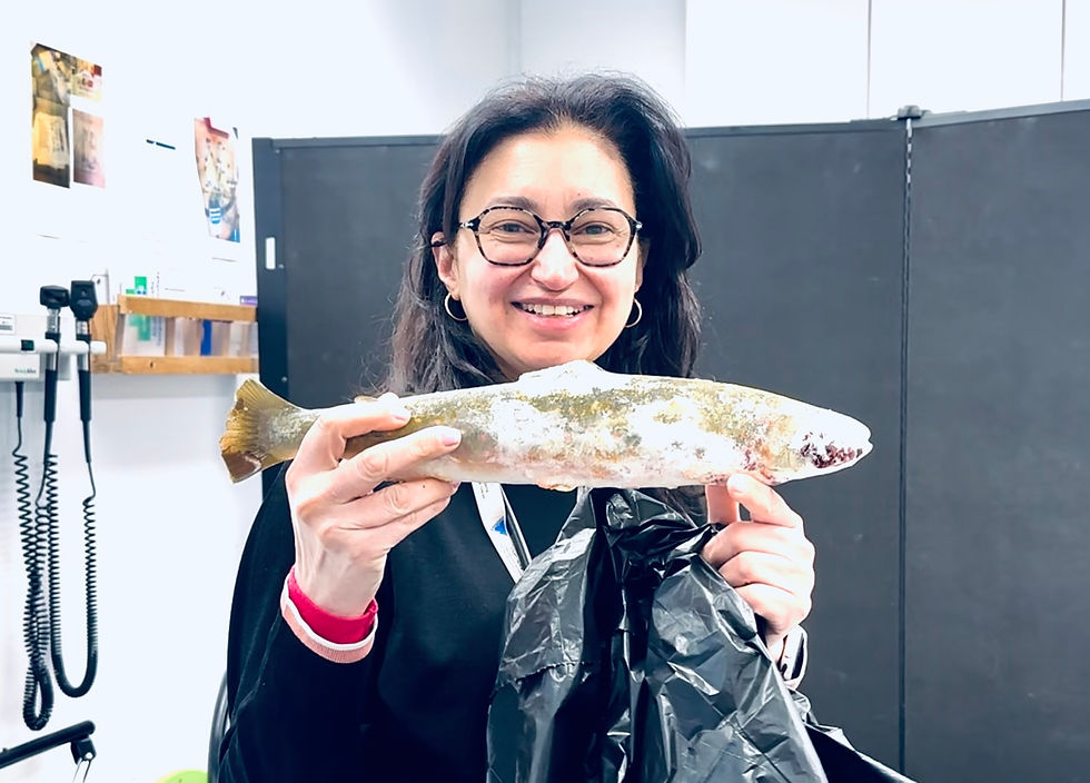 a smiling doctor holding a frozen Arctic char, standing in a medical clinic with dark cabinets and wall equipment behind.