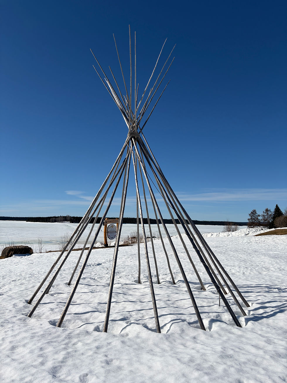 Wooden teepee structure set up on snow-covered ground by a frozen lake.