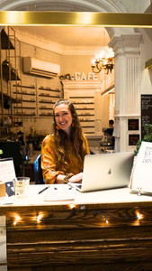 Danielle Klingelhofer sits at a laptop behind a welcome table at the front of a café, surrounded by books and string lights.