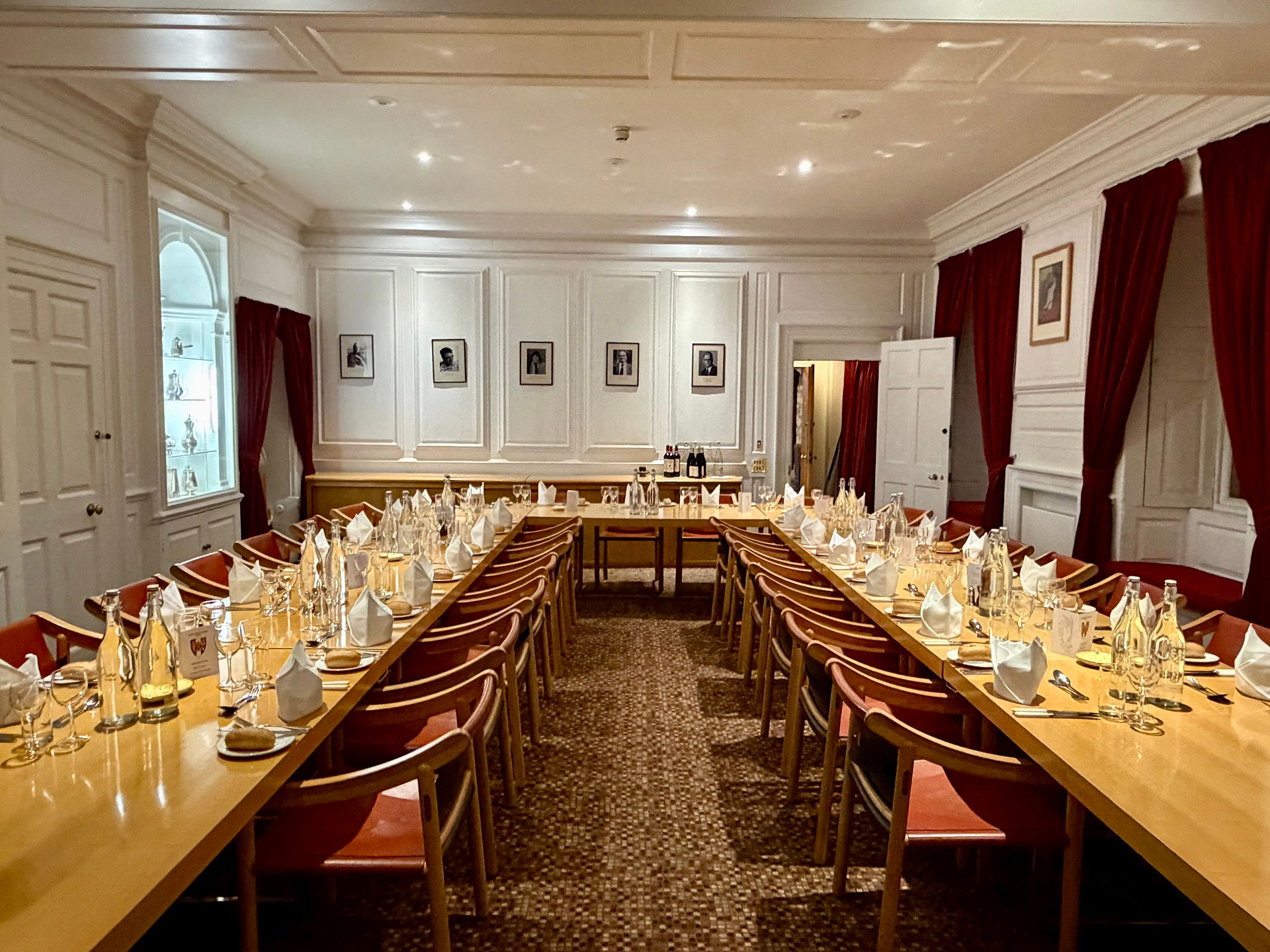 Long dining tables set with glassware and folded napkins inside a Corpus Christi college dining room with white panelled walls and red curtains.