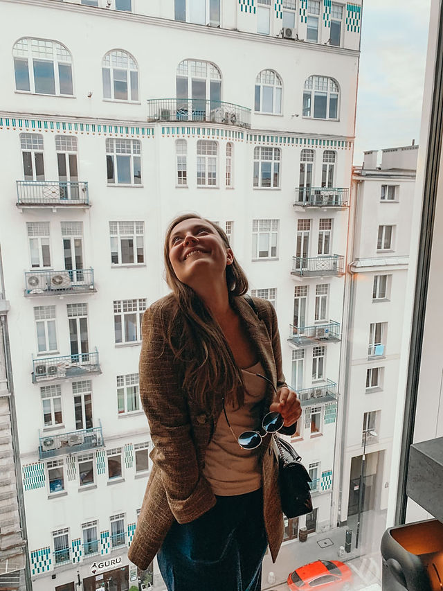 girl in brown jacket smiling in front of white european building