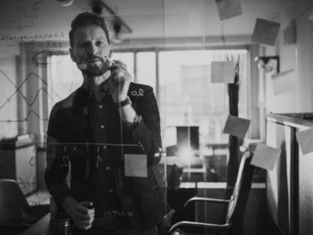 A professional in a modern office writes on a transparent glass board filled with graphs, equations, and sticky notes - capturing a moment of analytical focus and strategic problem-solving. Reflects the precision and clarity needed to build lasting demand in competitive markets.