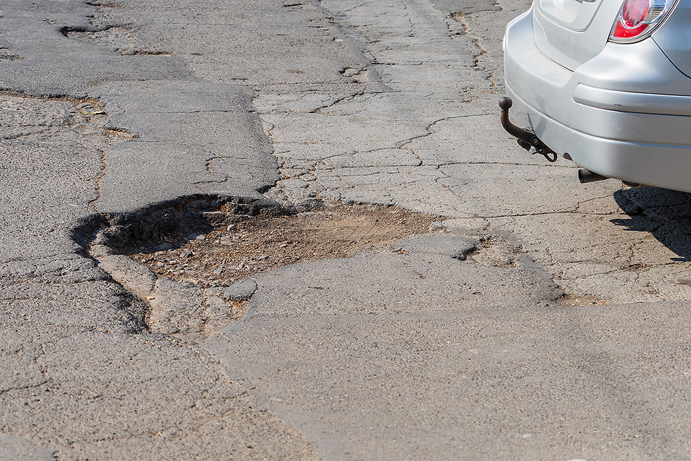 Cracked asphalt road with a large pothole beside the rear of a silver car with a tow hitch.