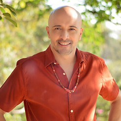 Pedro Lyion Astrólogo/Astrologia em meio a folhagens verdes usando uma camisa terracota e um colar de sementes
