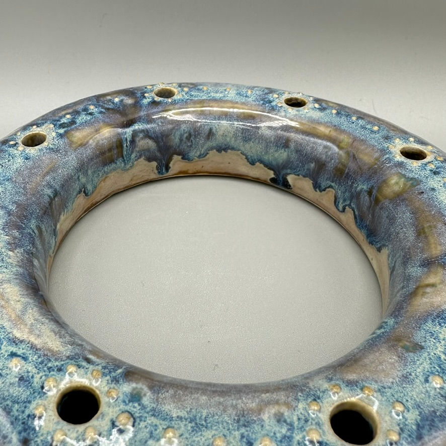 Round ceramic flower frog with a blue and gray glaze, featuring multiple holes for arranging flower stems.