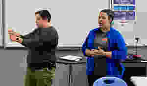 Two people standing at the front of a classroom, one is signing the other is speaking