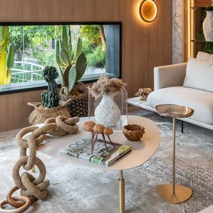 Sala de estar elegante com sofá branco, mesa de centro com revista OCA e colar de mesa, vista para jardim tropical.