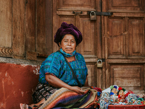 Woman in vibrant blue and purple clothing sits by wooden door, selling colorful handmade dolls and crafts from a basket, serene mood.
