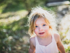 Blond toddler stands on sunlight dappled grass and smiles at camera. Image by Simon Rae on Unsplash.