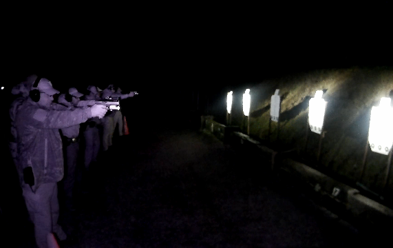 A line of people with handguns an flashlights practice shooting at targets seven metres away at night that are illuminated by the individual flashlights
