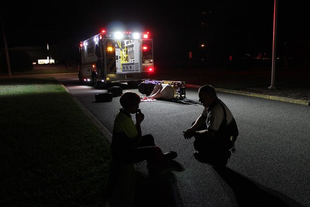 A paramedic comforts a person on a nighttime street while an ambulance with a knocked over gurney sits under lights in the background