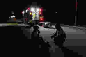 A paramedic comforts a person on a nighttime street while an ambulance with a knocked over gurney sits under lights in the background