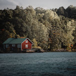 Haus am Seeufer: Ein rotes Haus steht malerisch am Ufer eines Sees, umgeben von herbstlichen Bäumen.
