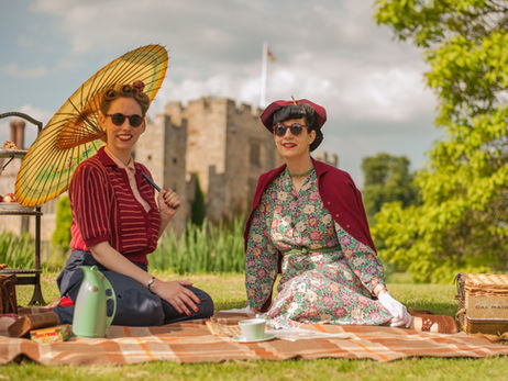Two women in vintage clothing have a picnic on a blanket, one holding a parasol. A castle is in the background, with a sunny, cheerful setting.